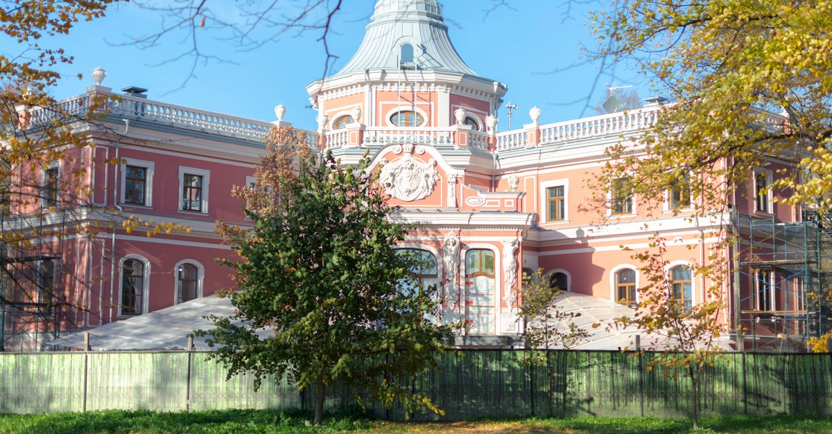 Elegant historic mansion with a pink facade set in a sunlit park, surrounded by autumn foliage.